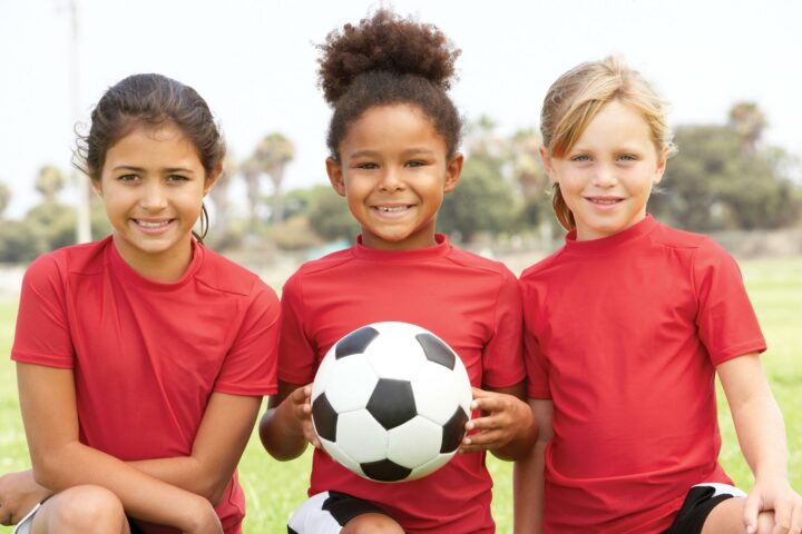 Three kids in red shirts on a field. The middle child is holding a soccer ball.