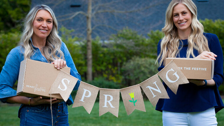 Two women hold up a 'spring' banner an ReadyFestive boxes.