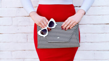 A woman holding a striped purse an white sunglasses, wearing red nails and a red pencil skirt.