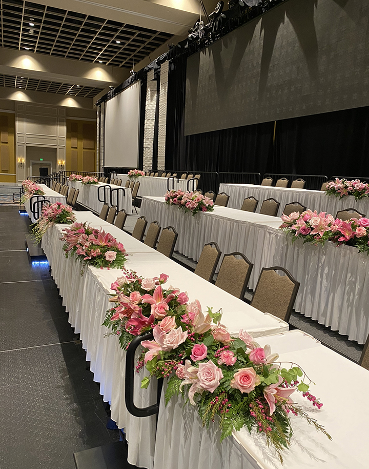 Three rows of white tables with pink and green flower bouquets and brown chairs.