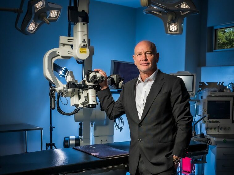 Dr. Masson wearing a black suit, standing next to medical equipment in a blue operation room.