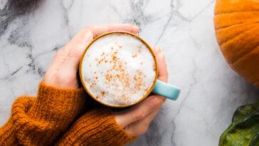 Autumn moody background with mug of pumpkin spice latte coffee and pumpkins on marble table. Flat lay in fall colors. Female hands in cozy sweater