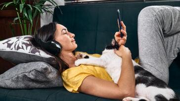 Woman listening to a true crime podcast with headphones lying embraced with her cat on the sofa.