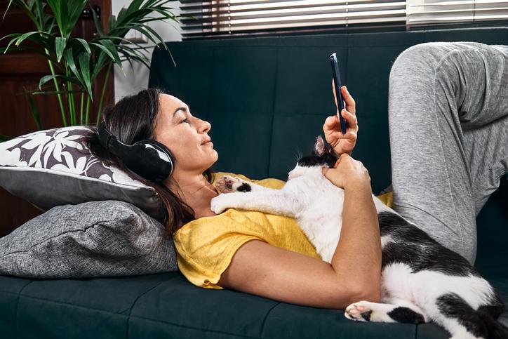 Woman listening to a true crime podcast with headphones lying embraced with her cat on the sofa.