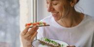 Smiling woman eating rye crisp bread with creamy vegetarian cheese tofu, cherry tomato and rucola micro greens, sitting at home and looking at window. Healthy food, gluten free, diet concept.