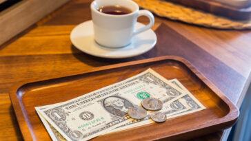 Dollar bills, coins and coffee on a table to represent the concept of tipping culture.