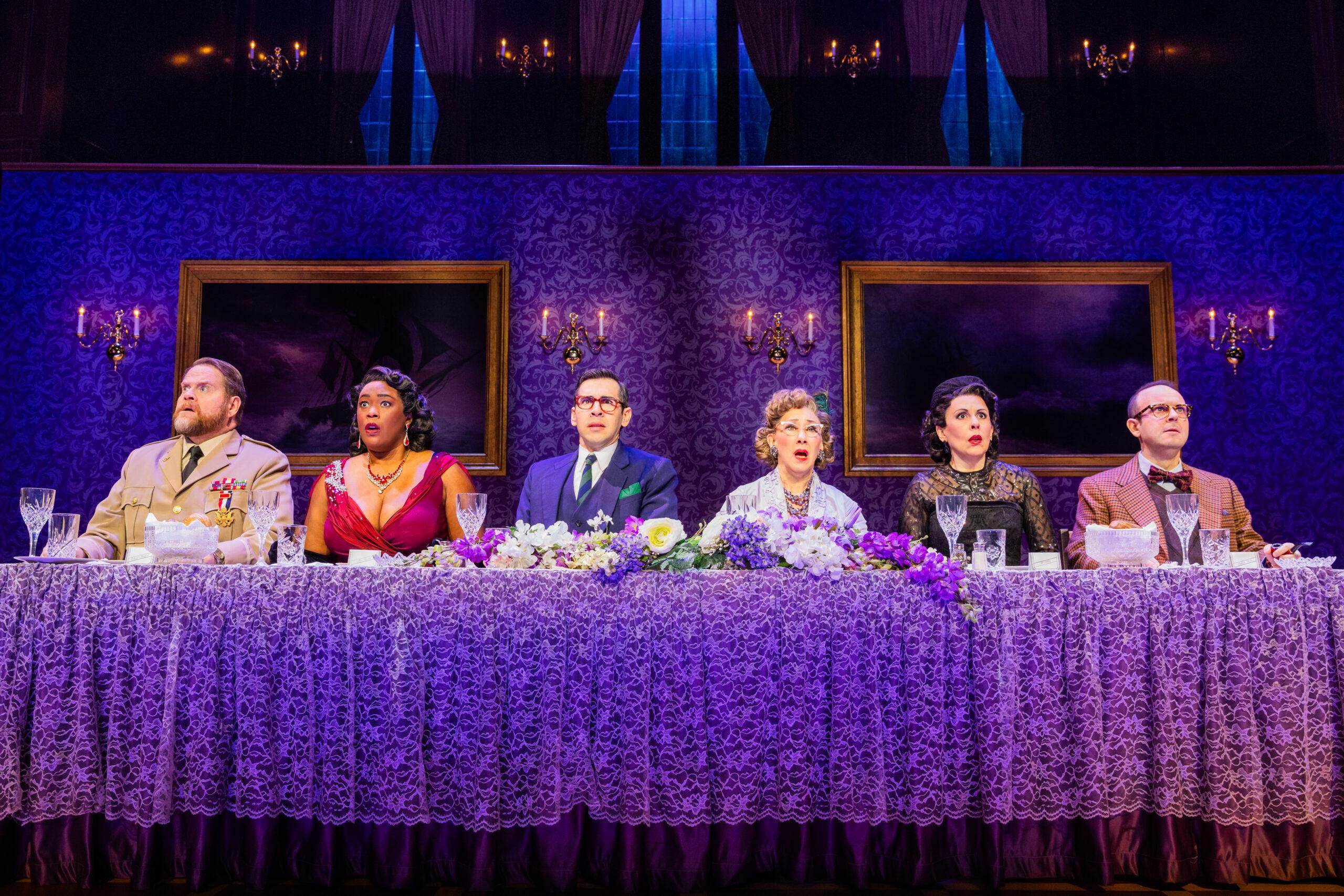 : Six actors in formal attire sit all on the same side behind a long fancy dinner table. Each actor is staring forward into the distance with a shocked and perplexed look. 