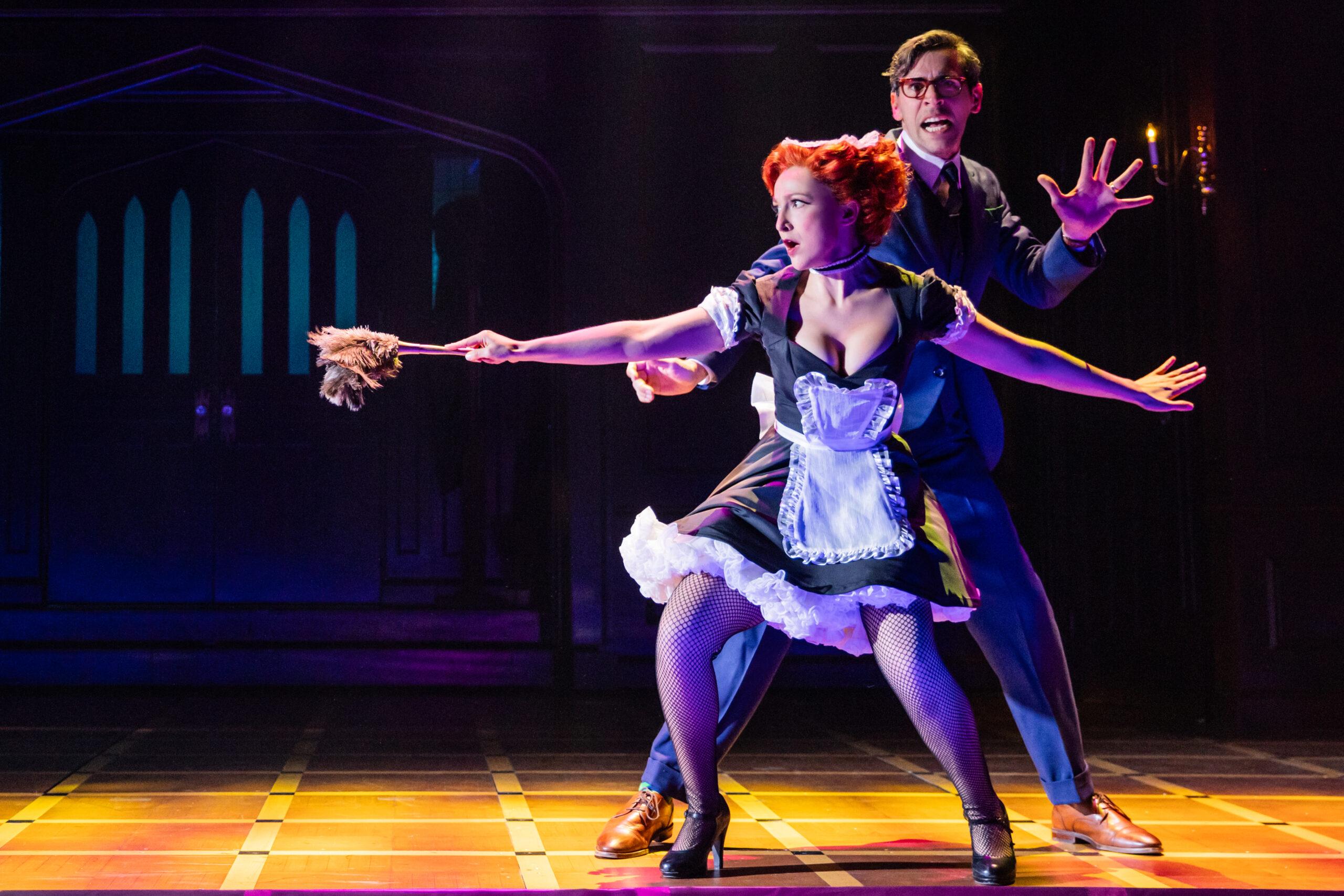 Two actors stand crouched on stage as they are on the lookout for something. A woman in a French maid’s uniform is slowly walking with a feather duster out in front of her as a weapon. A much taller man is behind her looking more frightened than her.